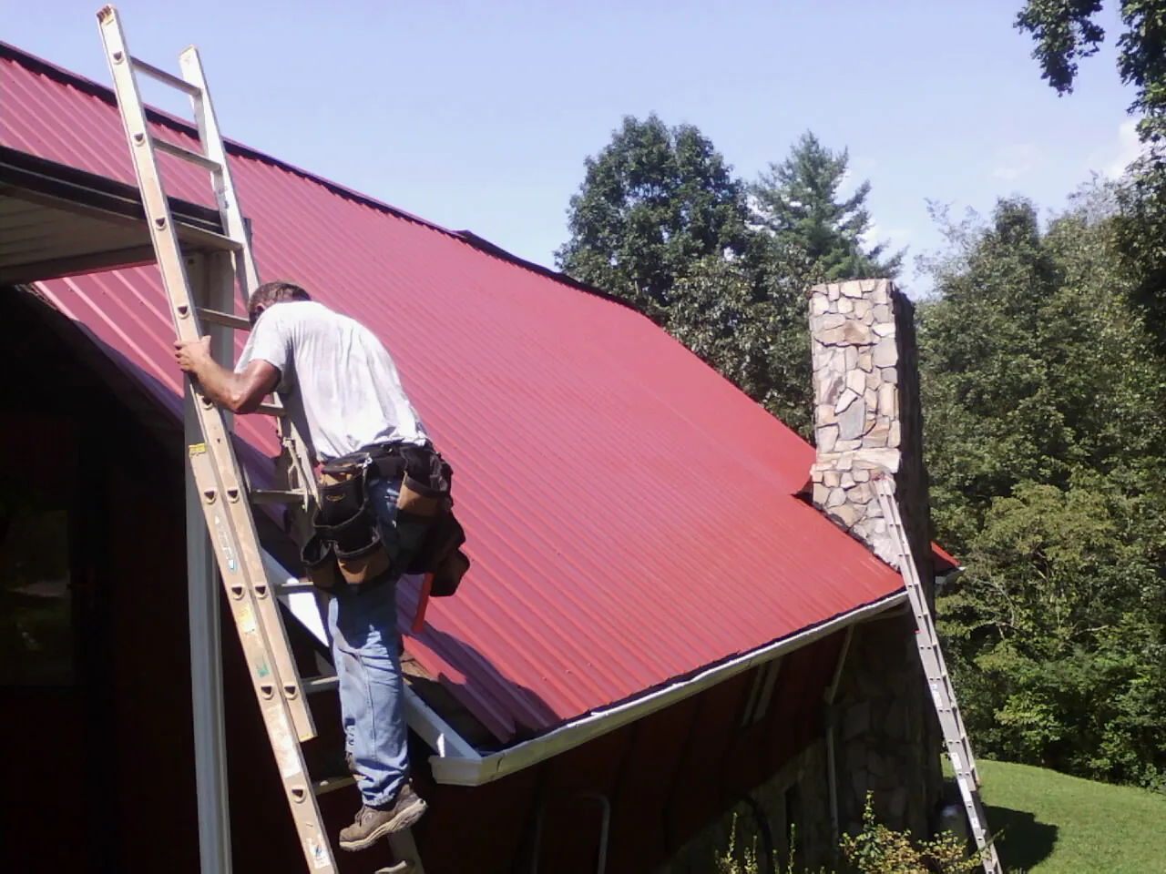 Roofer on a ladder beside a red metal roof and stone chimney — Soffit & Fascia Repair by Roofing Brothers in Indian Head